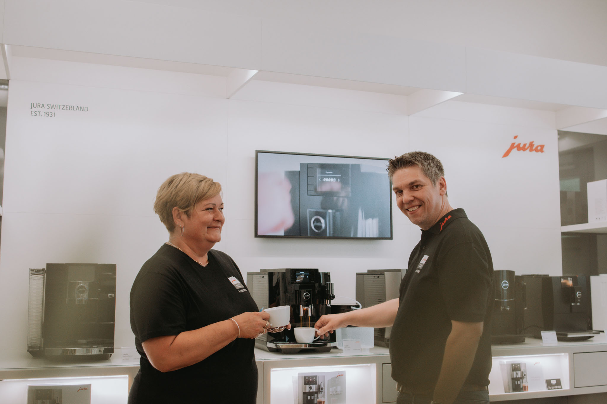 Hr. Geyer steht im Shop vor den Kaffeemaschinen und redet mit seiner Mitarbeiterin